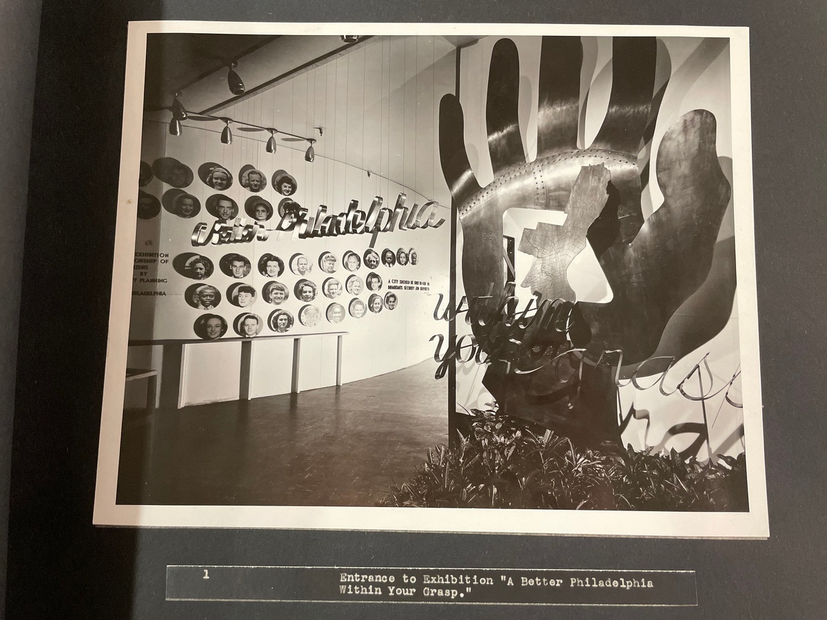 Photo of a large metal sculpture of a hand. The palm of the hand is cut out, and a metal map of Philadelphia is displayed in the cut-out. Thin strips of metal bent into a cursive font are suspended in the air in front of the hand. They spell out: A Better Philadelphia within your grasp