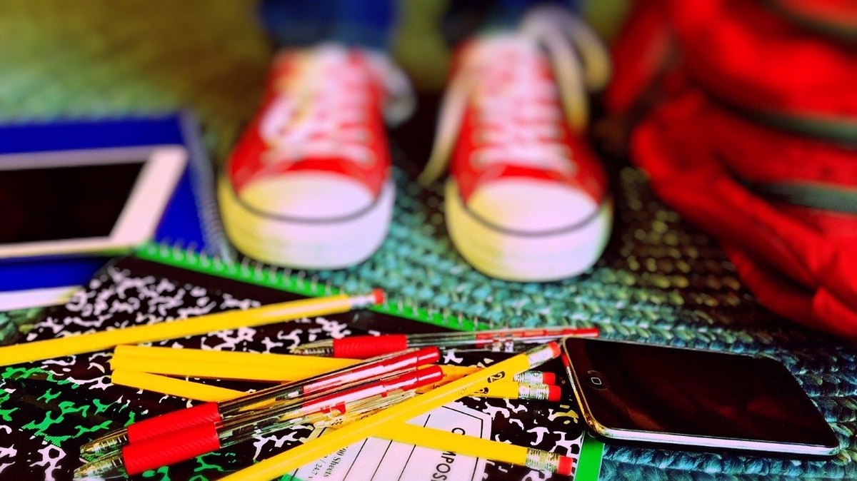 A student stands in red sneakers in front of a pile of pencils, notebooks, and school supplies. Are you ready to go back to school?