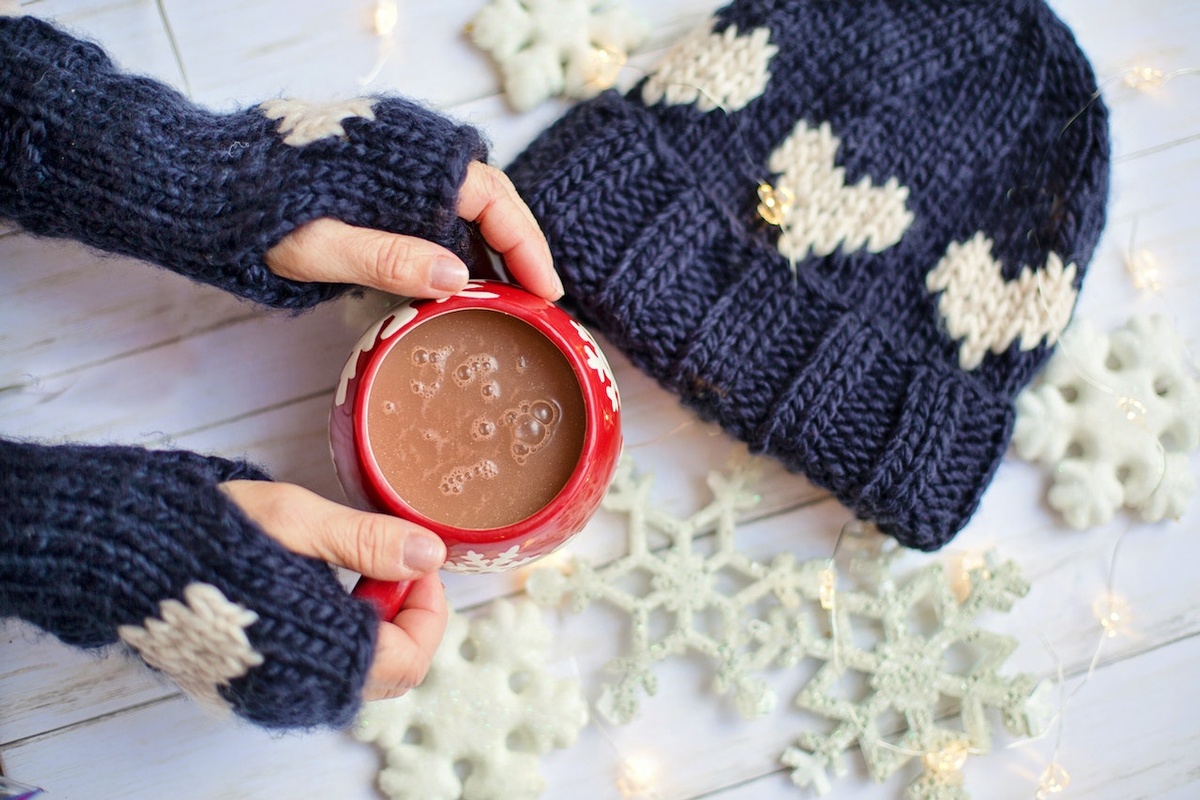 Two blue-mittened hands rest on a table, cradling a red mug of hot chocolate, beside a knitted blue hat with white hearts.