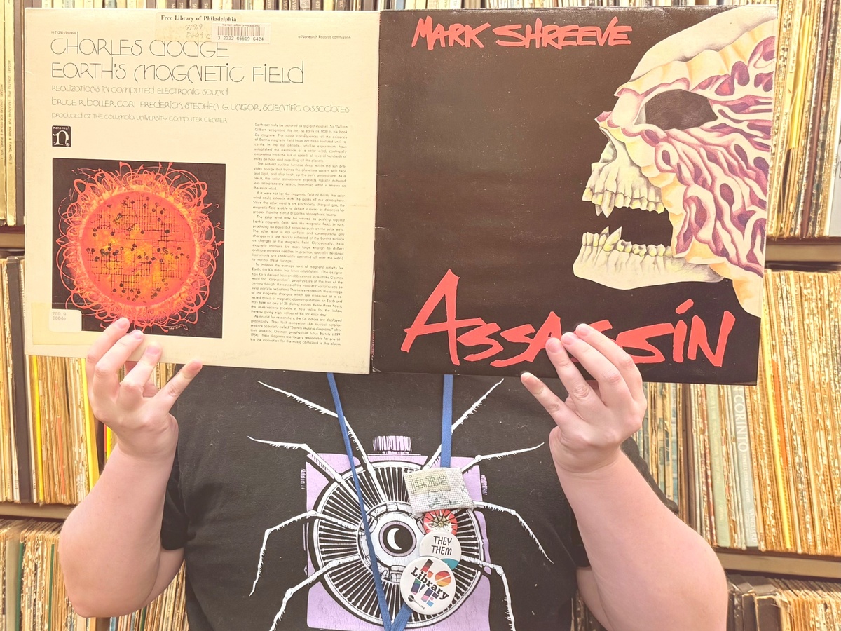 Librarian posting with vinyl record sleeve covering their face with library stacks of vinyl records behind them